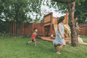 A blog about summer activities Indiana foster families can enjoy. Depicts a woman, a man, and a young boy running and playing together in a grassy backyard near a wooden play structure with a slide.